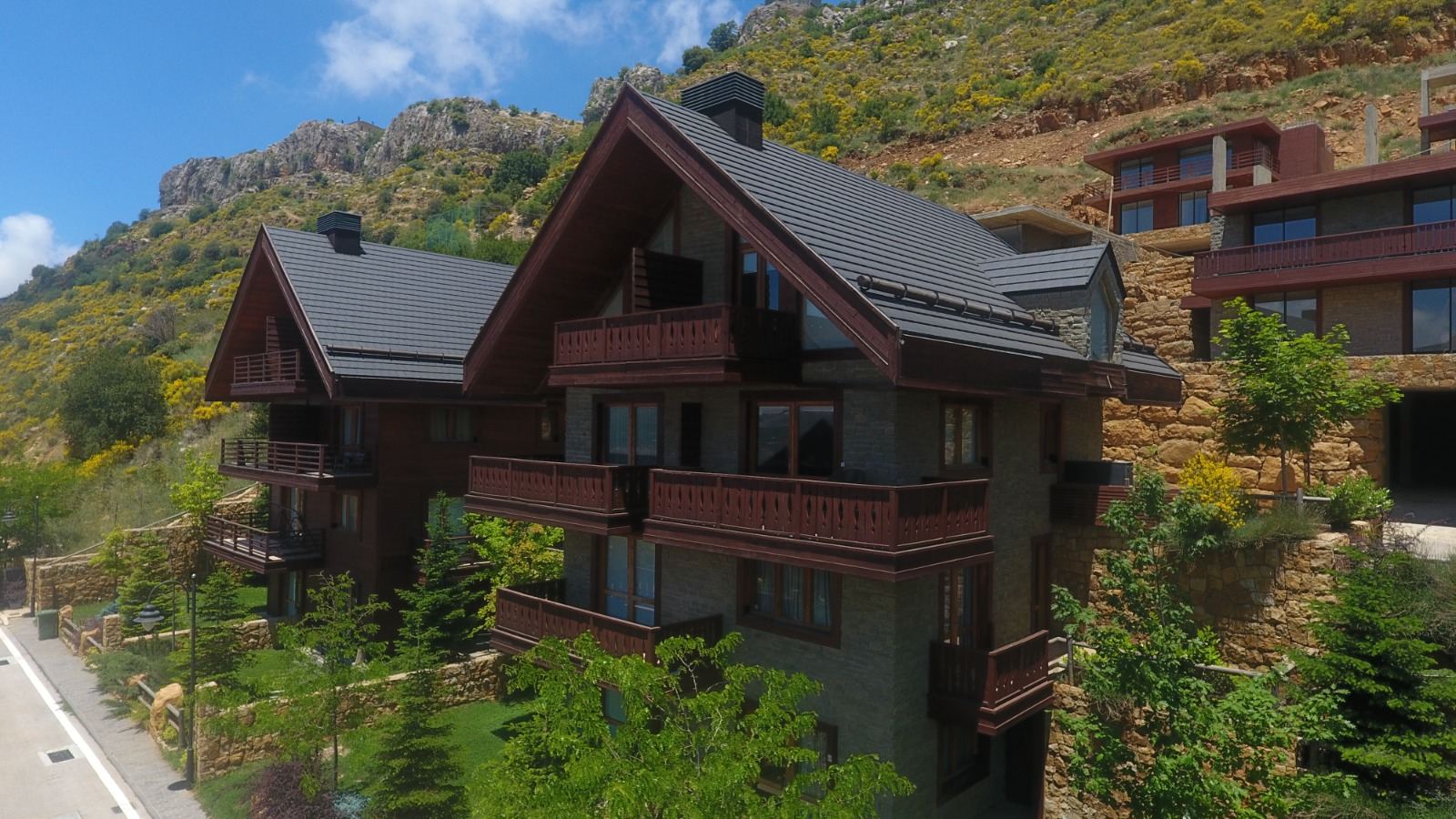 A dark-timber and grey-stone chalet with deep balconies and a steep slate-grey pitched roof, seen slightly from above against a rocky ridge covered in yellow flowering broom under a deep blue sky.