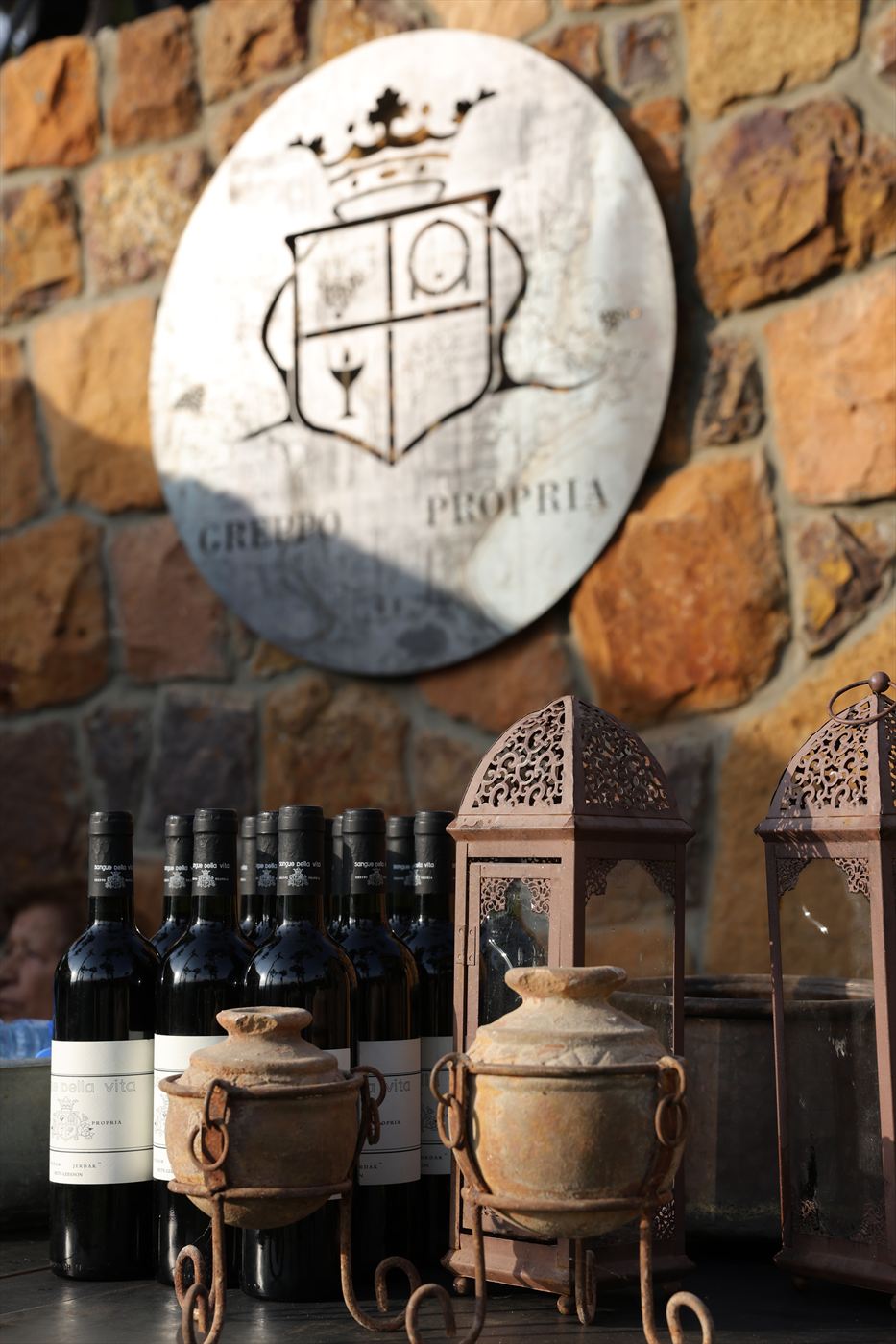 A pale marble disc engraved with the Greppo Propria coat of arms, mounted on a wall of rough golden-brown rubble stone, with a row of dark red-wine bottles standing below it between two terracotta urns in rusted iron stands.